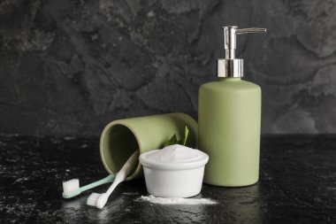 Yapraklı bir kase diş tozu, tutacaklı fırçalar ve siyah arka planda duran sabun makinesi.