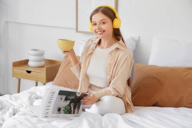Kulaklıklı, kahve fincanı ve iç tasarım dergisiyle evde yatakta oturan genç bir kadın.