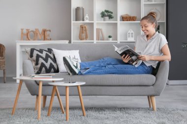 Evdeki kanepede iç tasarım dergisi okuyan genç bir kadın.