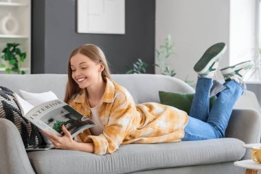 Evdeki kanepede iç tasarım dergisi okuyan genç bir kadın.