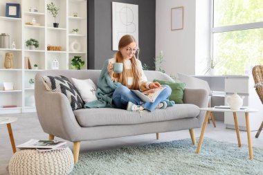 Evdeki kanepede kahve fincanı okuyan genç bir kadın.