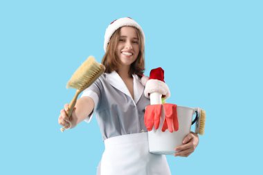 Mavi arka planda Noel Baba şapkası ve temizlik malzemeleri olan genç oda hizmetçisi.