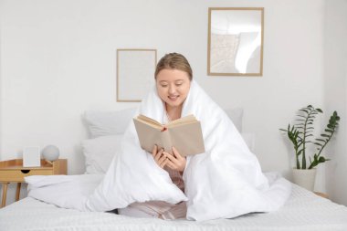 Güzel, genç, mutlu bir kadın battaniye içinde evde kitap okuyor.