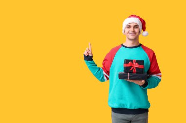 Handsome young man in Santa hat holding Christmas gifts and pointing at something on yellow background clipart