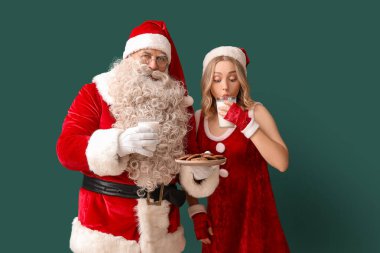 Noel Baba ve güzel kadın yeşil arka planda kurabiyeli süt içiyorlar.