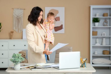 Evde doğum izni belgelerini okurken dizüstü bilgisayarla çalışan sevimli küçük bebek ve genç anne.