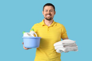 Mavi arka planda bir yığın temiz elbisesi ve çamaşır deterjanı olan genç bir adam.