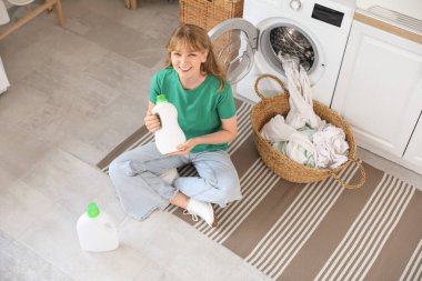 Young woman with laundry detergent sitting near washing machine at home clipart