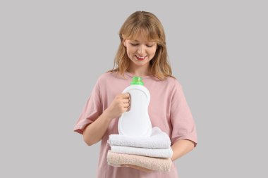 Çamaşır deterjanı ve gri arka planda bir yığın temiz havlusu olan genç bir kadın.