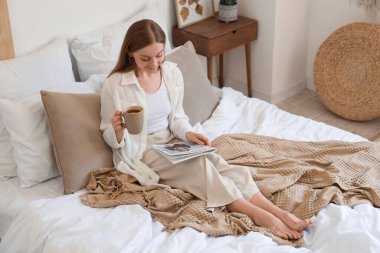 Elinde çay ve dergi olan genç bir kadın evde yatağında oturuyor.
