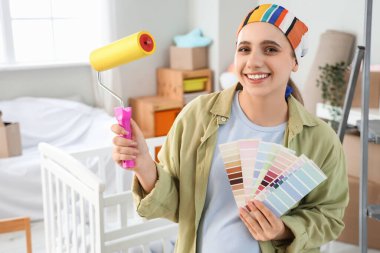 Çocuk odasındaki tamirat sırasında boya silindirli ve renk paletli hamile bir kadın.