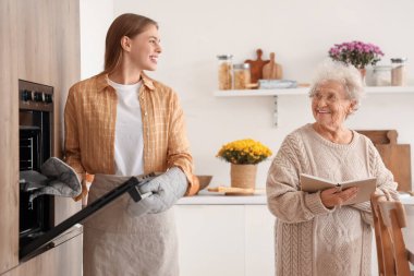 Fırından fırın tepsisi alan genç bir kadın ve mutfakta yemek tarifi kitabıyla büyükannesi.