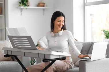 Evdeki kanepede dizüstü bilgisayarı olan genç bir kadın sentezleyici oynuyor.