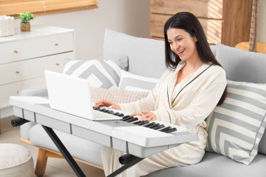 Pijamalı genç bir kadın evdeki kanepede sentezleyici oynuyor.