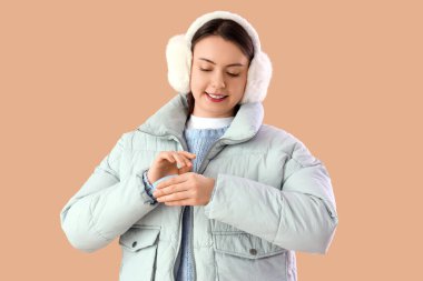 Kışın giyinmiş genç bir kadın bej arka planda el kremi sürüyor.