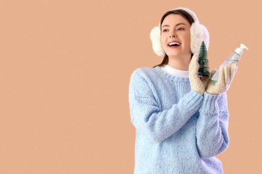 Cilt bakımı ürünü ve bej arka planda oyuncak Noel ağacıyla kışın giyinmiş genç bir kadın.