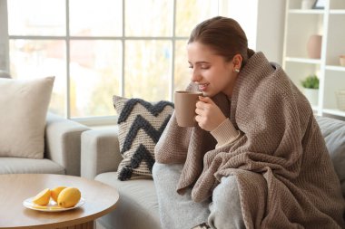 Ekoseli gülümseyen genç kadın evdeki kanepede limon çayı içiyor.
