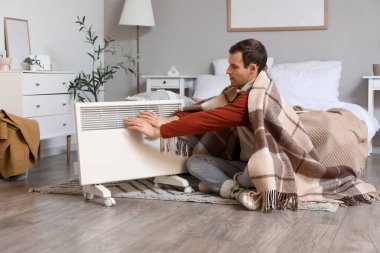 Yatak odasındaki elektrikli konteyner ısıtıcısının yanında ekoseli elleri olan genç bir adam.