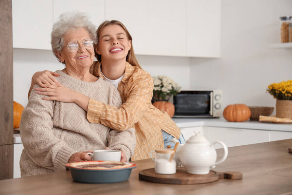 Молодая женщина обнимает свою бабушку с чаем и вкусным пирогом на столе на кухне