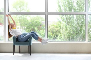 Geniş pencerenin yanındaki koltukta uzanan genç bir kadın.