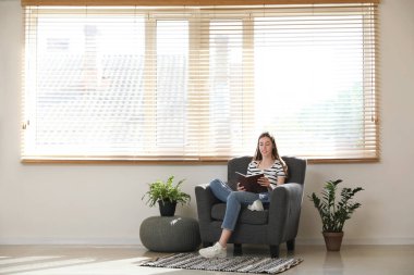 Oda bitkilerinin yanındaki koltukta oturmuş kitap okuyan genç bir kadın.