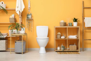 Beyaz klozet, sehpa ve turuncu duvarın yanında raf ünitesi olan şık bir tuvalet.