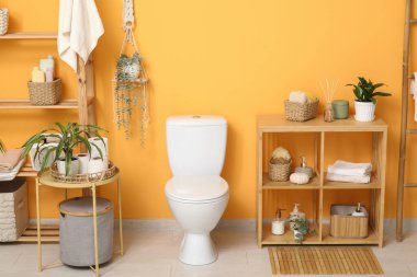 Beyaz klozet, sehpa ve turuncu duvarın yanında raf ünitesi olan şık bir tuvalet.