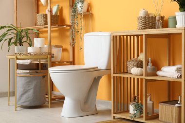 Beyaz klozet, sehpa ve turuncu duvarın yanında raf ünitesi olan şık bir tuvalet.