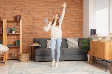 Kulaklıklı, tablet bilgisayarlı, evde dans eden genç bir kadın.