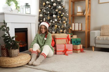 Noel Baba şapkalı, sıcak çoraplı genç bir kadın Noel arifesinde şöminenin yanında oturuyor.