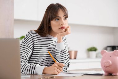 Düşünceli genç bayan mutfaktaki masada para sayıyor.