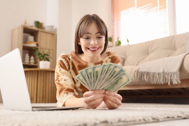 Evde yerde yatan dolar banknotları ve dizüstü bilgisayarı olan genç bir kadın.