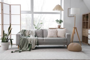 Gri kanepe, lambalar ve geniş pencereli aydınlık oturma odasının içi.