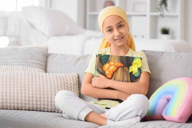 Little girl after chemotherapy with yellow ribbon and book sitting on sofa at hospital. Childhood cancer awareness concept clipart