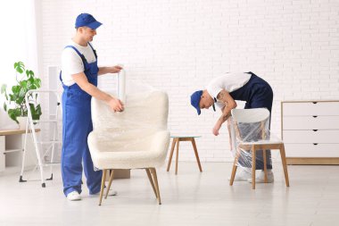 Erkek yükleyiciler odada streç filmli sandalyeleri paketliyor.