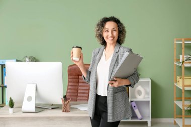 Olgun iş kadını, elinde klasör ve işyerinin yanında modern bilgisayarla şık bir ofiste kahve tutuyor.