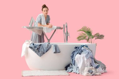 Pembe arka planda ütü masası, çamaşır ve küveti olan yorgun genç bir kadın.