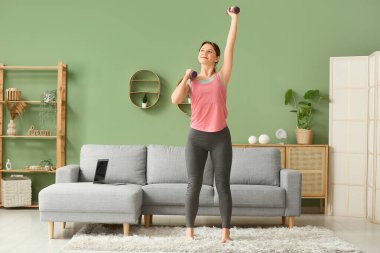 Oturma odasında dambıllarla antrenman yapan güzel bir genç kadın.
