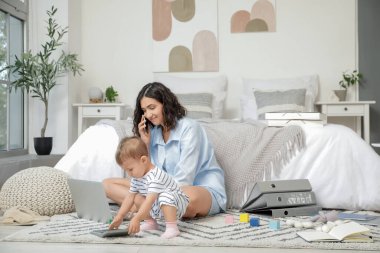 Çalışan anne cep telefonuyla konuşuyor ve küçük bebeği hesap makinesiyle yatak odasında.