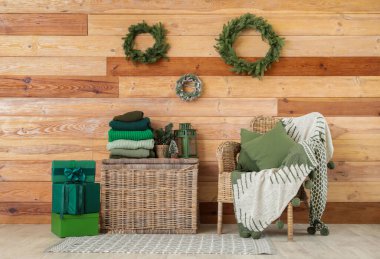 Wicker armchair, basket with stack of winter clothes and Christmas wreathes on wooden wall clipart