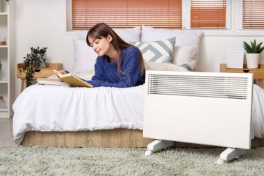Yatak odasındaki elektrikli ısıtıcının yanında kitap okuyan genç bir kadın.