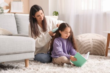 Genç anne evde oturmuş kitap okuyan küçük kızının saçlarını tarıyor.