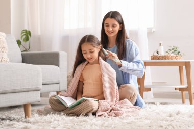 Genç anne evde oturmuş kitap okuyan küçük kızının saçlarını tarıyor.