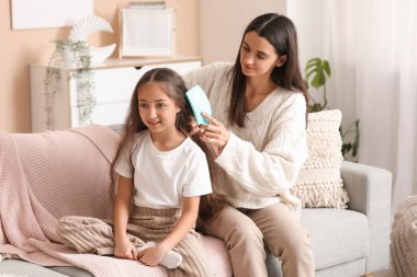 Genç anne, evdeki kanepede otururken küçük kızının saçını tarıyor.