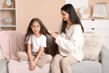 Genç anne, evdeki kanepede otururken küçük kızının saçını tarıyor.