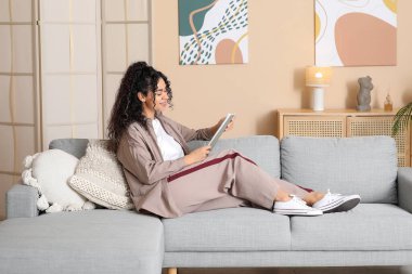 Güzel bir Afro-Amerikalı kadın, şık bej gömlekli, dergisi evde, kanepede oturuyor.