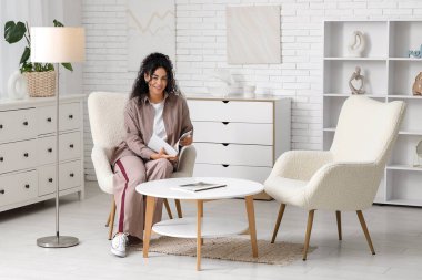 Güzel bir Afro-Amerikalı kadın şık bej gömlekli ve dergisi evde koltukta oturuyor.