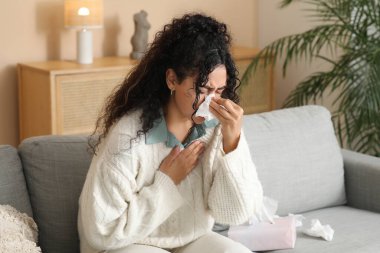 Mendilin içinde hapşıran ve evdeki kanepede oturan genç Afro-Amerikalı kadın. Alerji konsepti