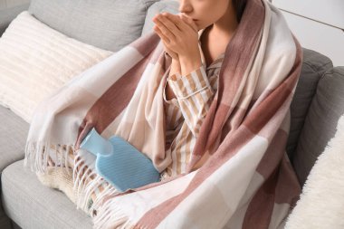 Pijamalı, sıcak su şişesi ve battaniyeli genç bir kadın evdeki kanepede oturuyor.