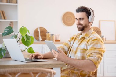 Kulaklıklı yakışıklı adam mutfaktaki masada dizüstü bilgisayar ve tablet kullanıyor.